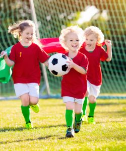 Kids play football on outdoor field. Portugal team fans with national flag. Children score a goal at soccer game. Child in Portuguese jersey and cleats kicking ball. Fan celebrating victory at pitch.